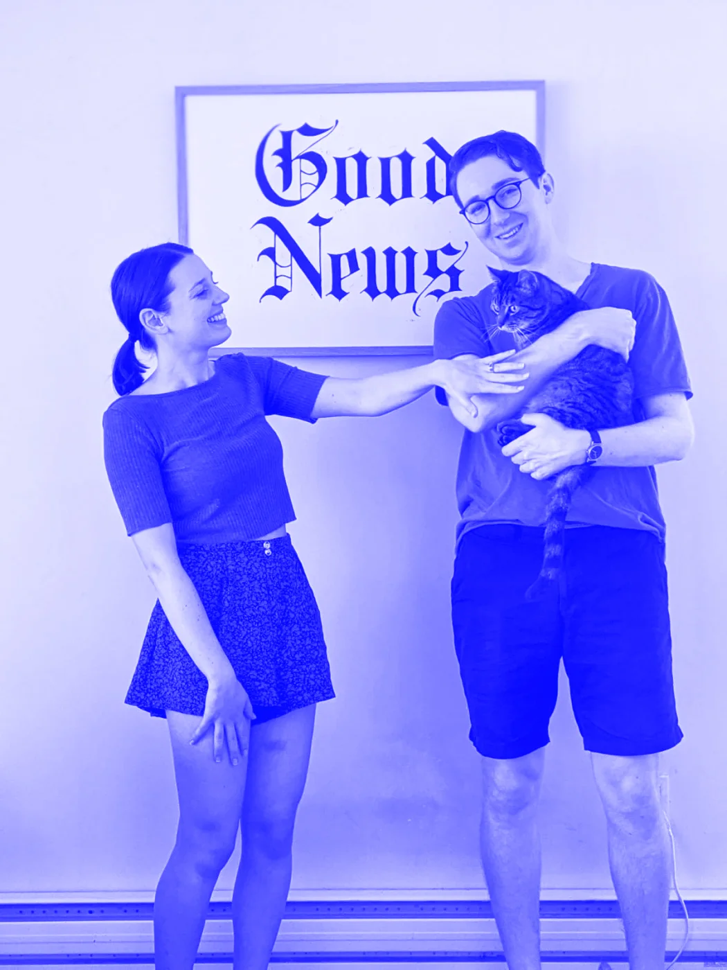 Patrick and Jess with their cat standing in front of a poster that reads 'Good News'. Jess has her engagement ring on.